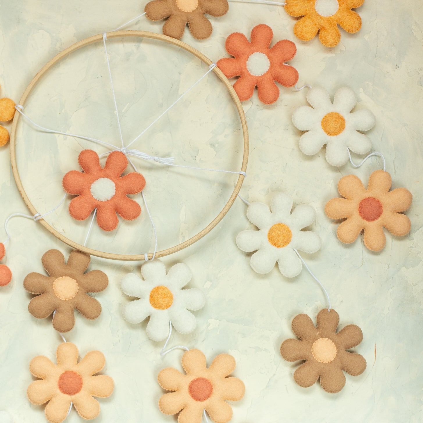 A handmade baby mobile hanging from a wooden arm, featuring several tiers of felt daisies in shades of terracotta, peach, tan, and white suspended from a light wooden hoop.