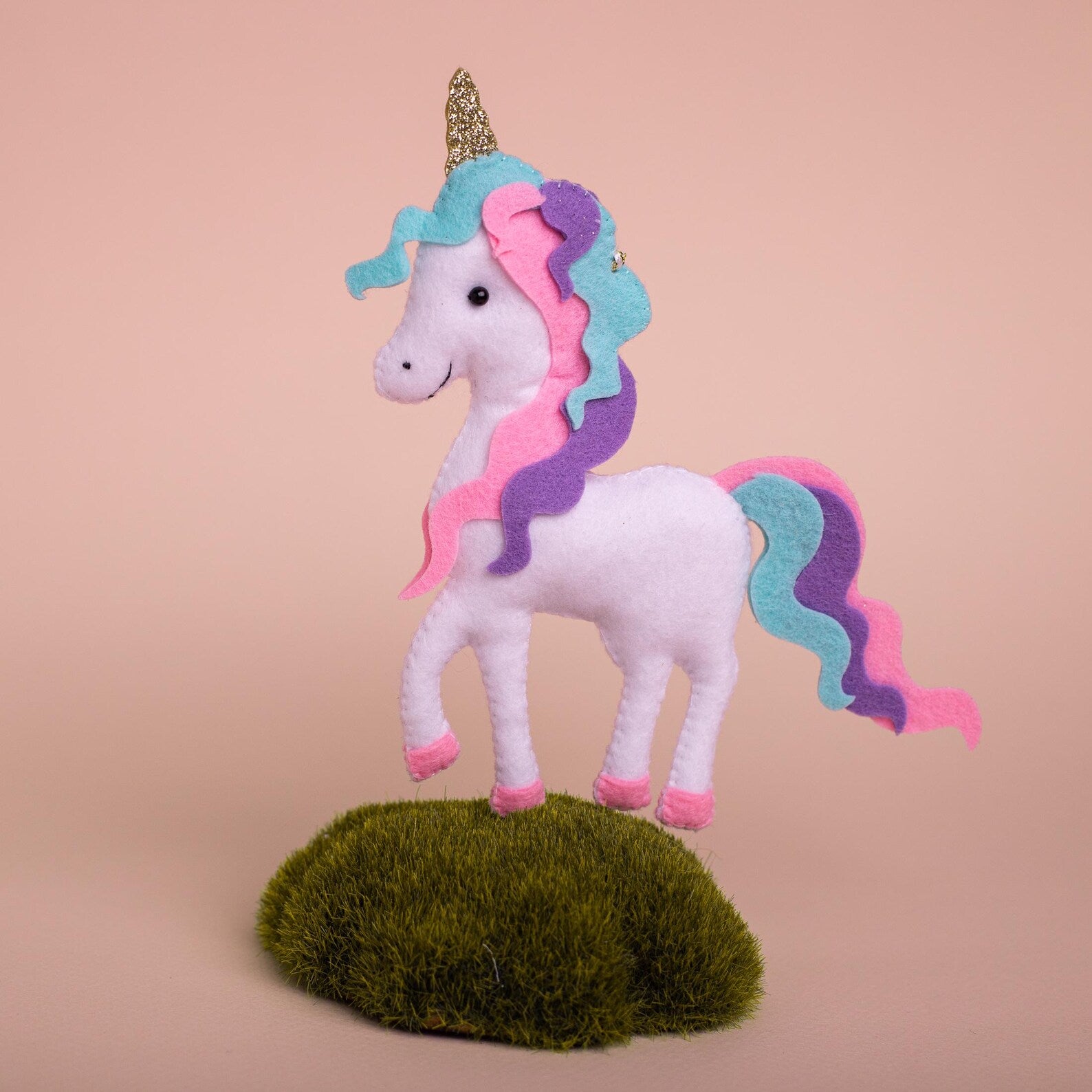A handmade white felt unicorn ornament with a wavy pastel mane and tail in pink, purple, and teal, a sparkly gold horn, and pink hooves, with a gold loop for hanging.