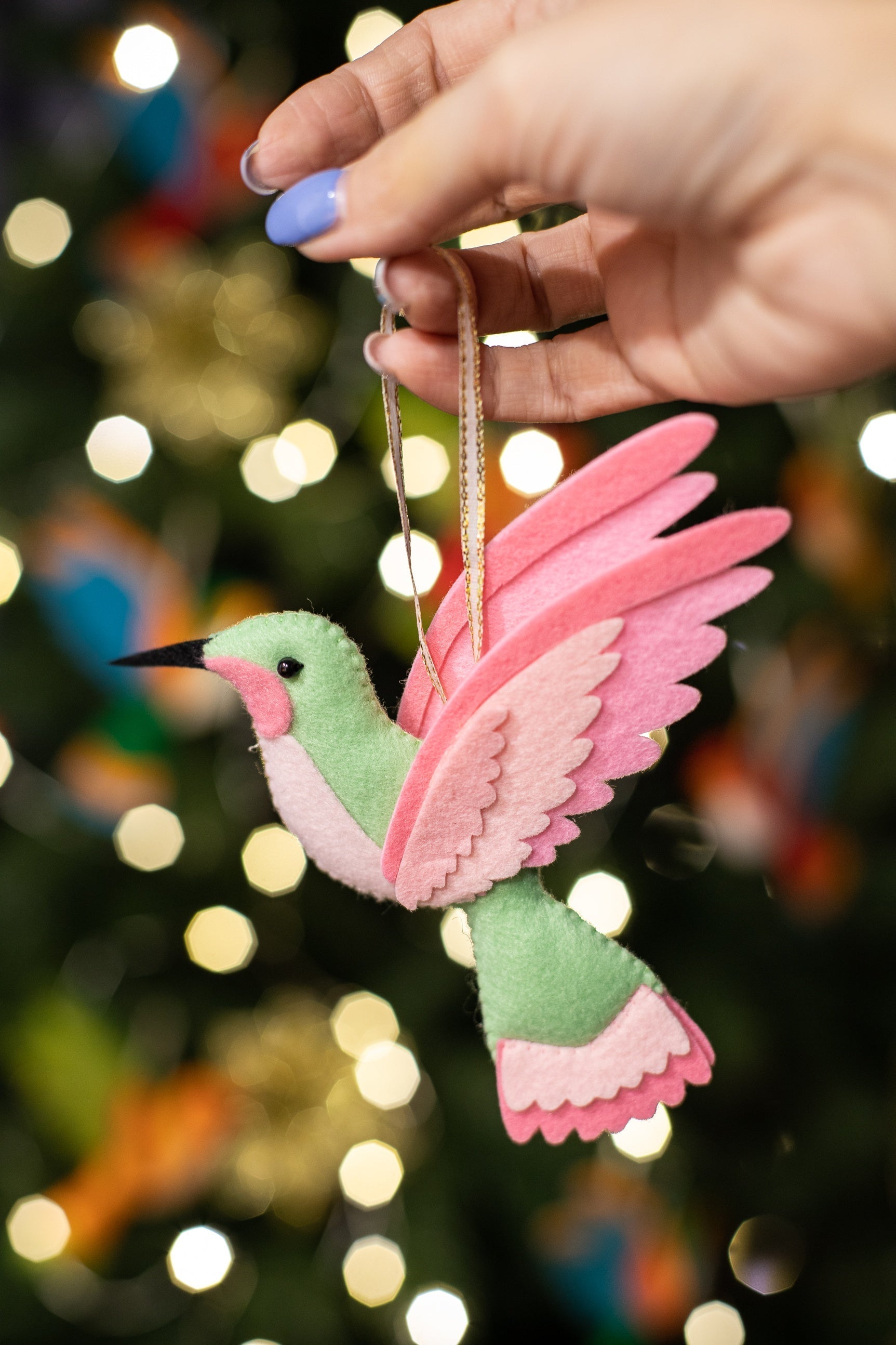 A hand holds a handmade felt hummingbird ornament. The bird is mint green with a black beak. Its wings are a blend of pink and light pink. The background is a blurred Christmas tree with glowing festive lights.