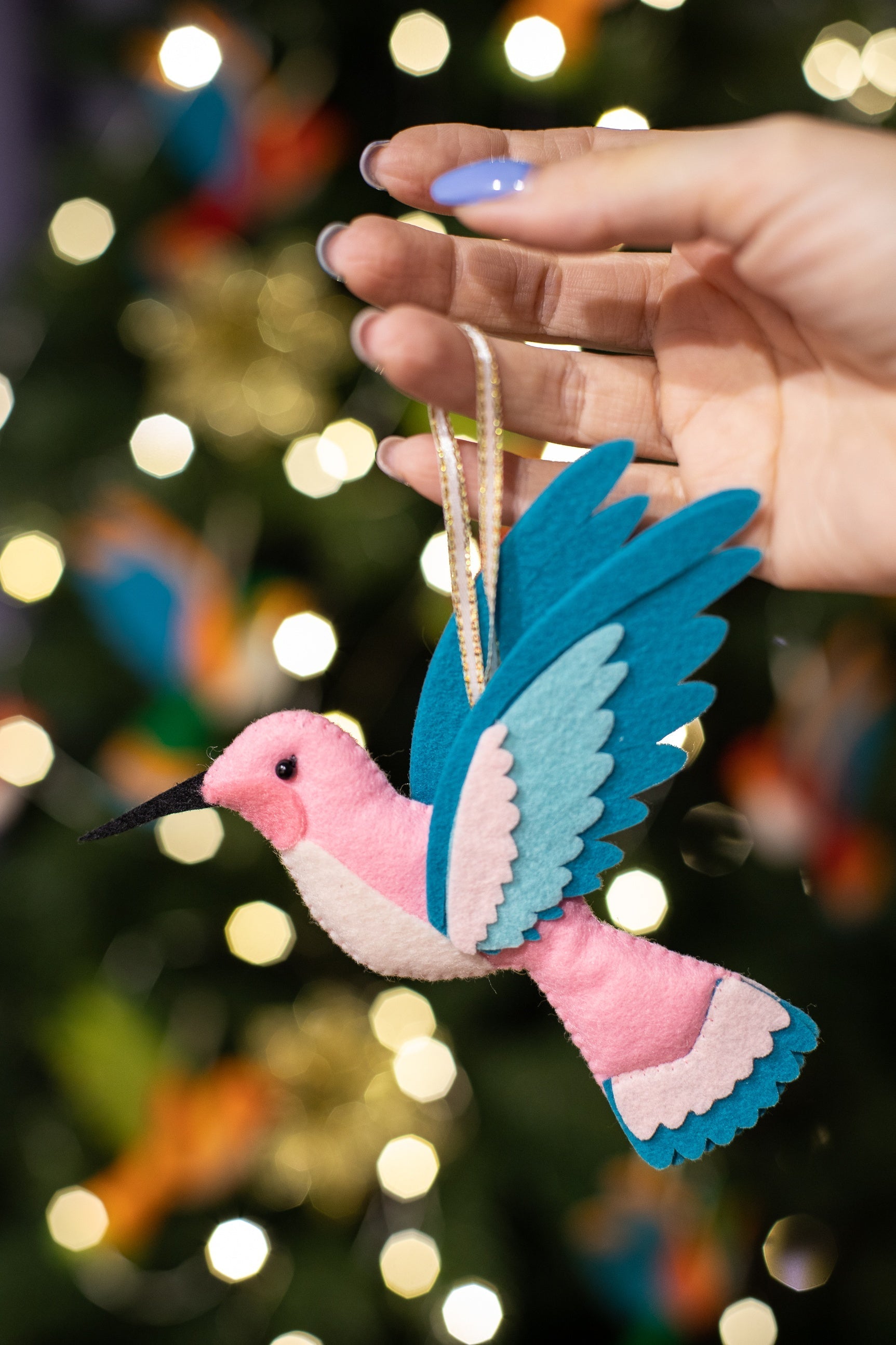 A handmade pink felt hummingbird ornament with a white belly, light blue wings, and a black beak