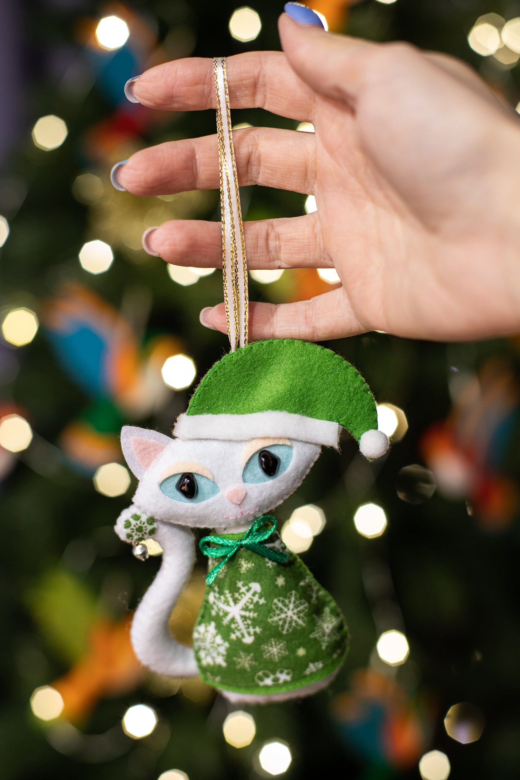 White felt cat ornaments with light blue eyes and curled tails. The cat wears a green hat with a white pom-pom and a green dress with white snowflakes.