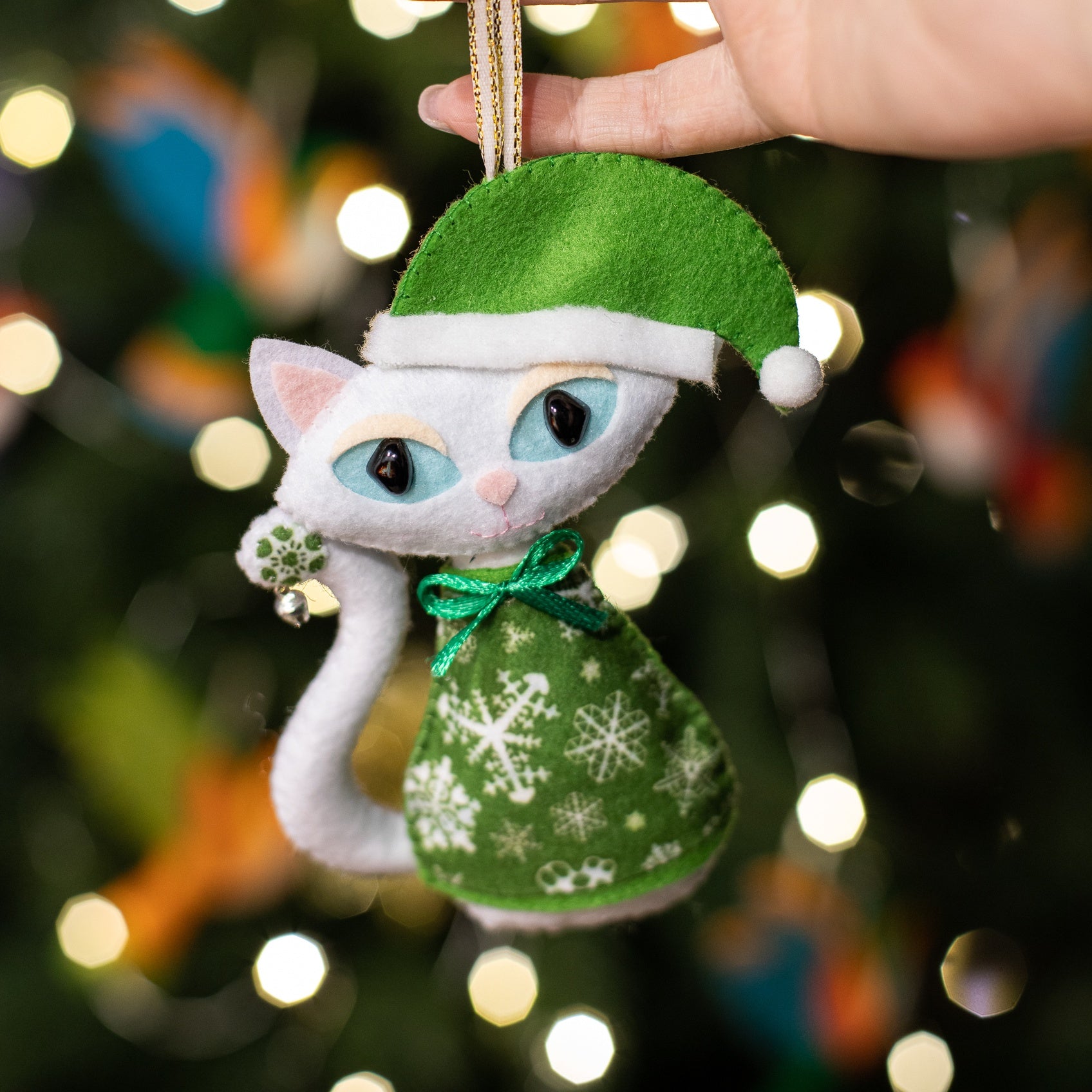 White felt cat ornaments with light blue eyes and curled tails. The cat wears a green hat with a white pom-pom and a green dress with white snowflakes.