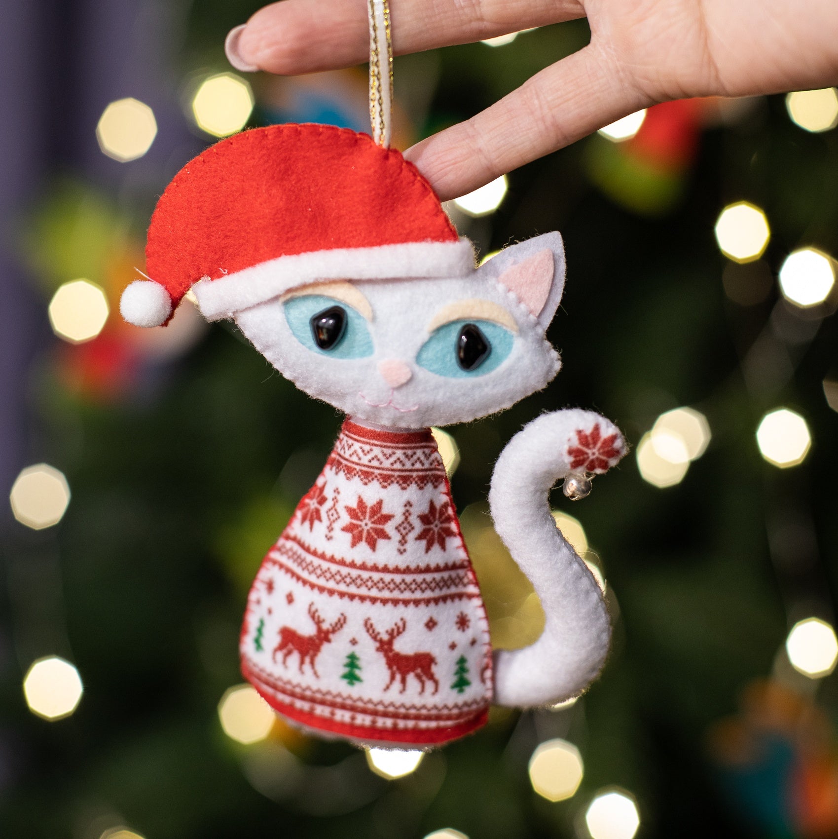 Felt cat ornaments with light blue eyes and curled tails. The cat wears a red Santa hat with a white pom-pom and a red sweater with white deer and snowflake patterns.