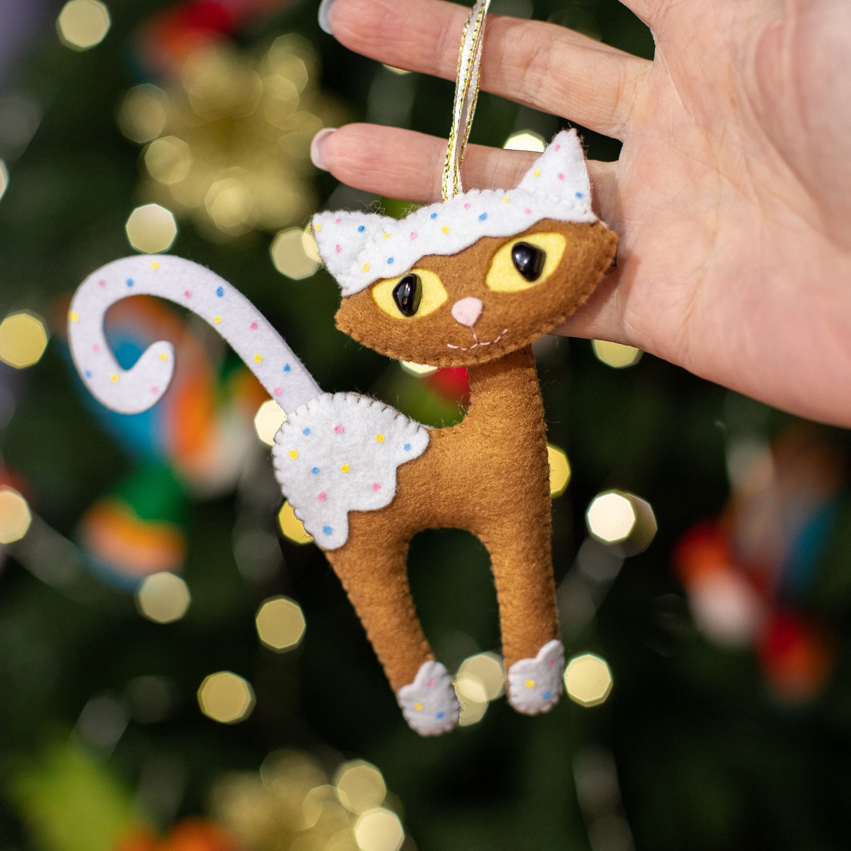 A handcrafted brown felt cat with white paws, chest, and a white tail. The cat has large yellow eyes and white felt pieces with multi-colored polka dots resembling icing on its head, paws and tail.