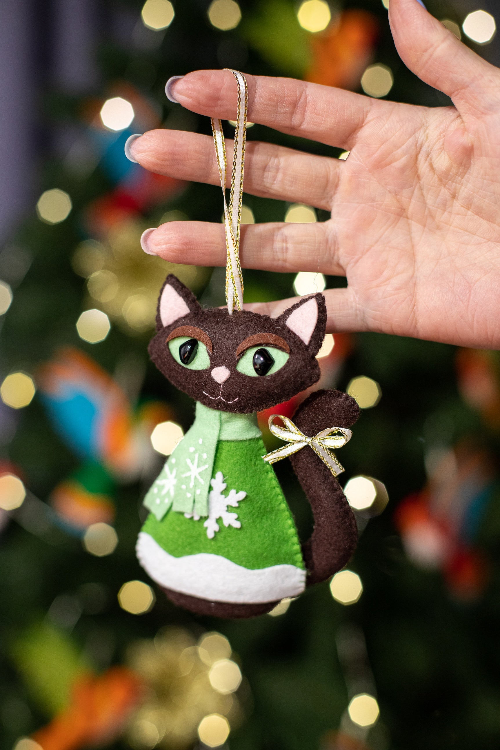 Brown felt cat Christmas ornament wearing a mint green scarf and green sweater with snowflake design. The cat has brown felt body,  black eyes, and a small gold bow on its tail. 