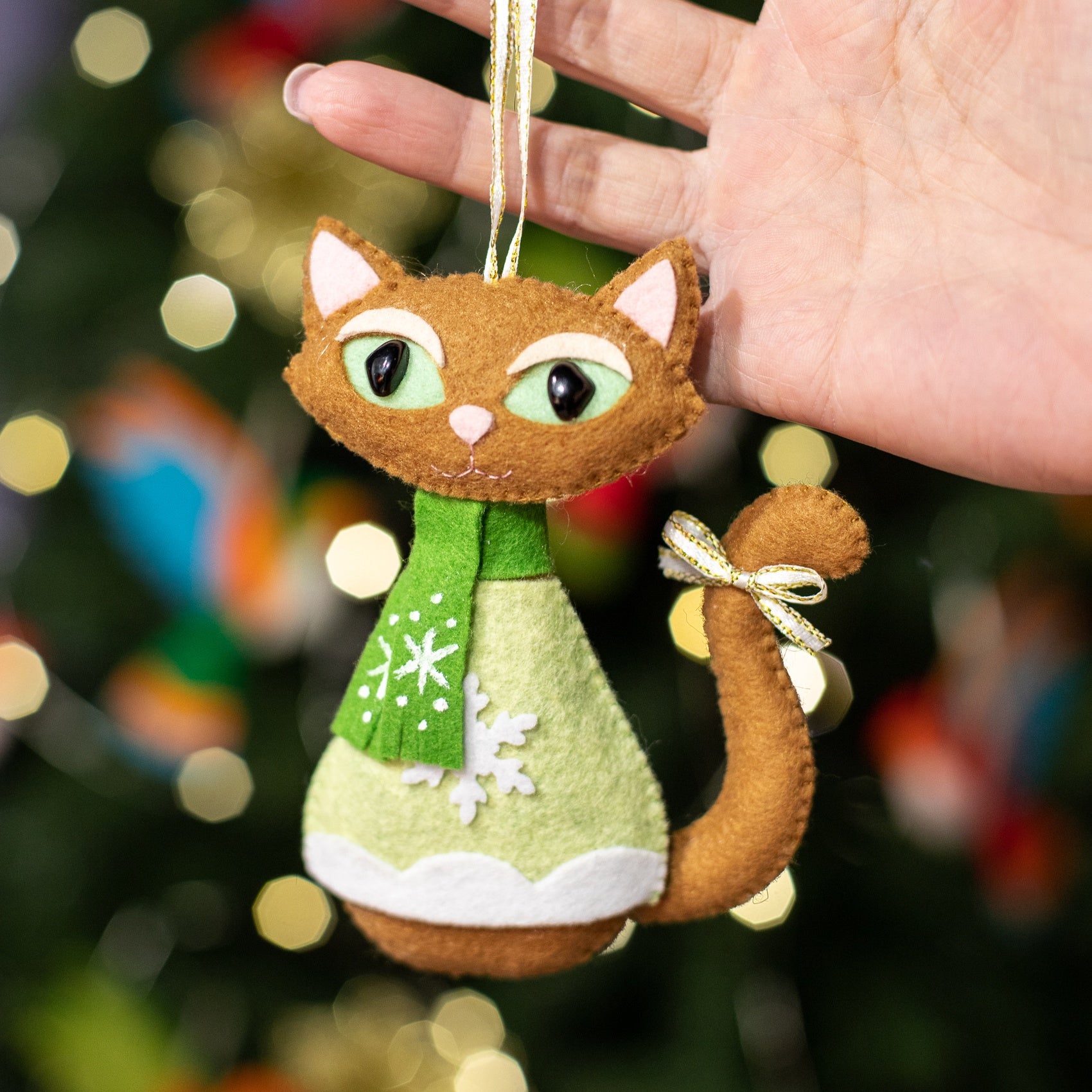 A felt Christmas cat ornament. The cat is brown with green and white snowflake patterned scarf. It has a light brown tail and gold bow. The cat is wearing a light green sweater with white trim at the bottom.