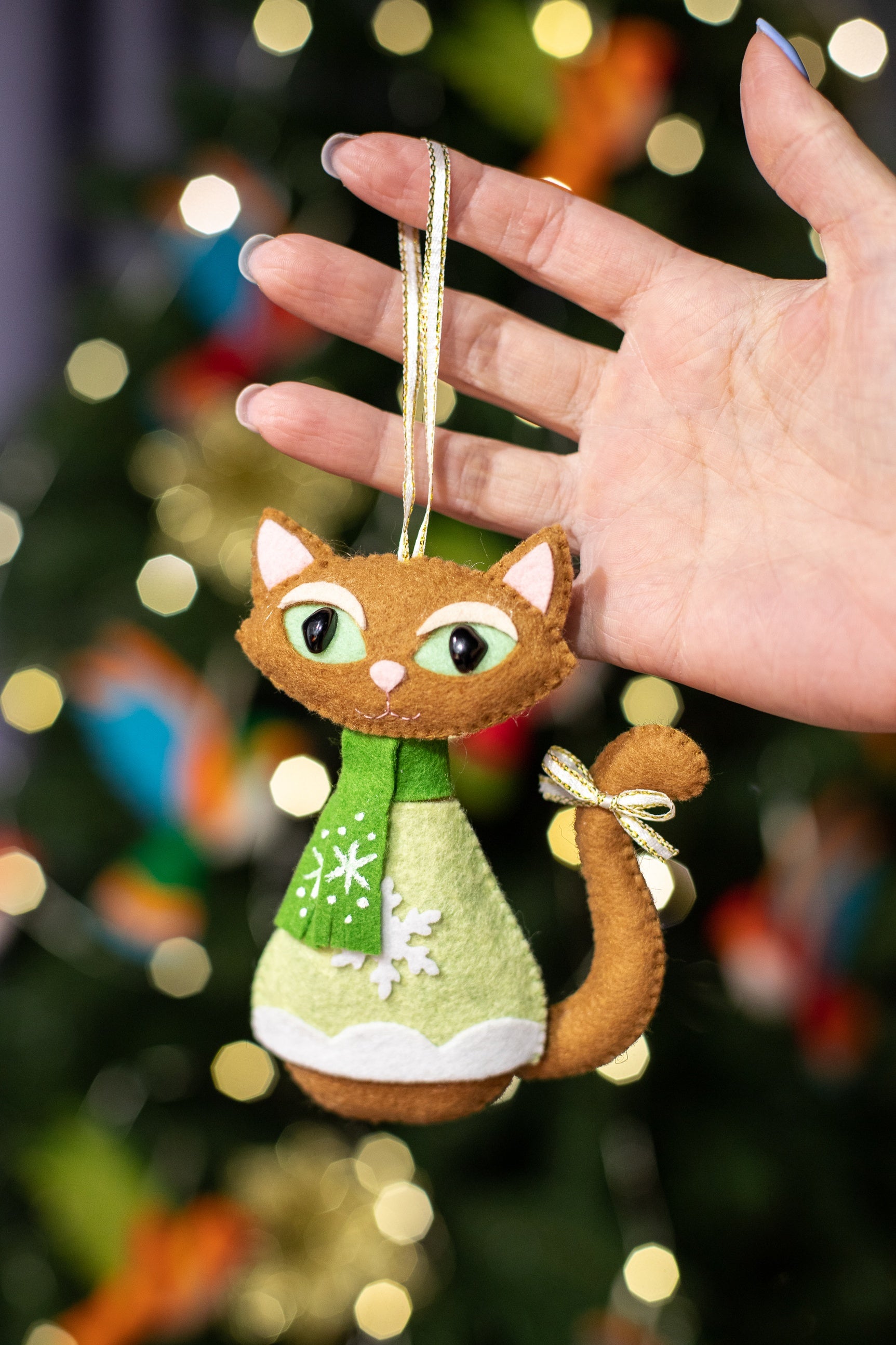 A felt Christmas cat ornament. The cat is brown with green and white snowflake patterned scarf. It has a light brown tail and gold bow. The cat is wearing a light green sweater with white trim at the bottom.