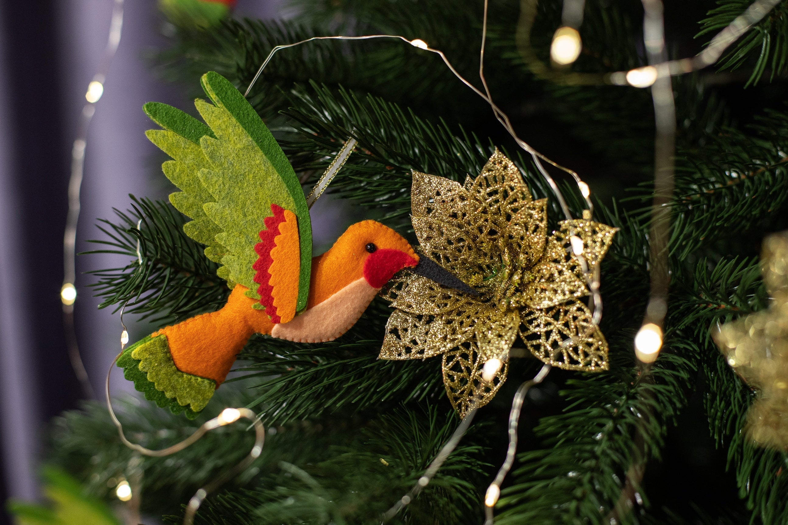 A handmade orange felt hummingbird ornament with a biege belly, green wings, and a black beak