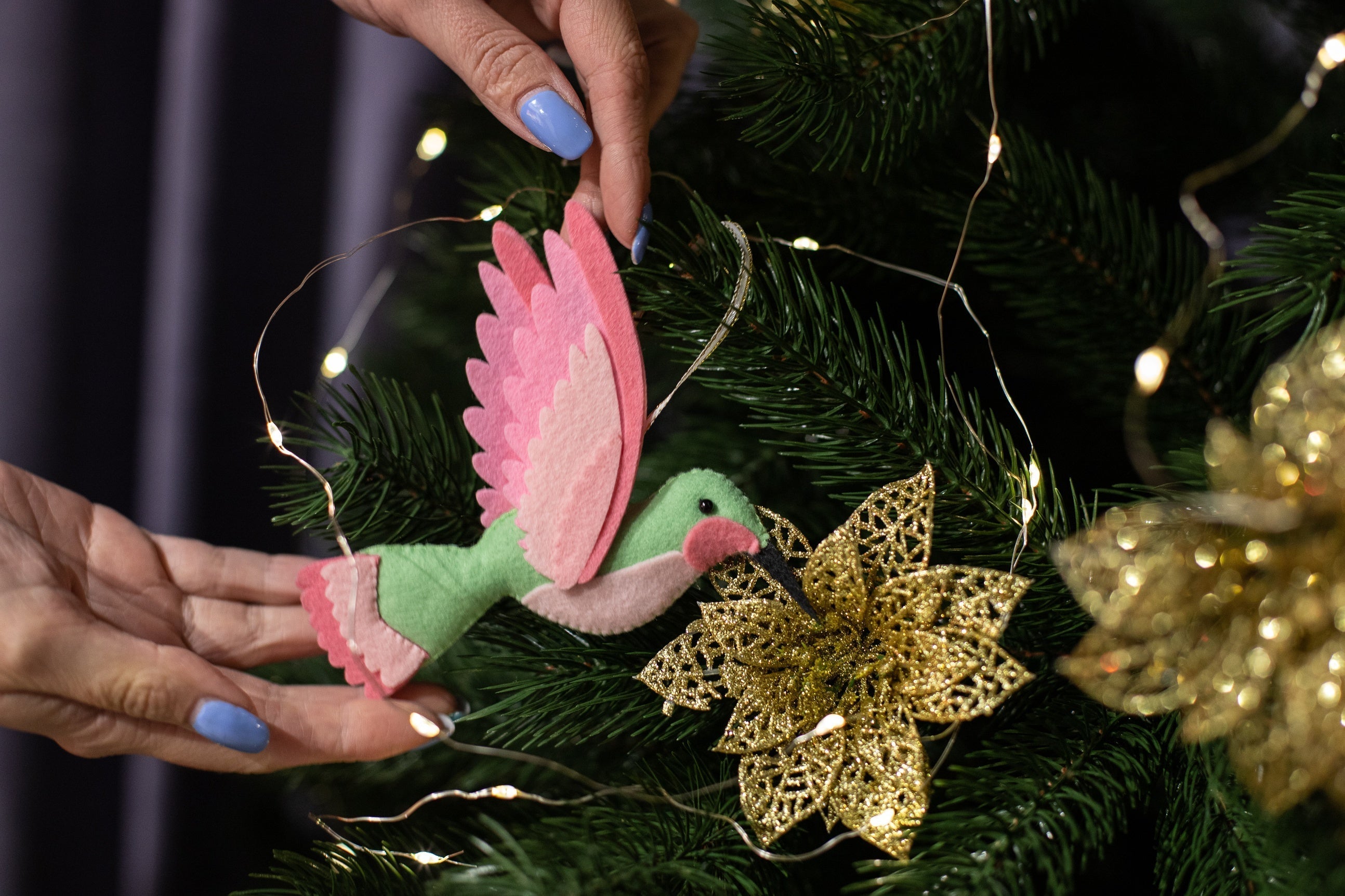 Handmade felt hummingbird ornament. The bird is mint green with a black beak. Its wings are a blend of pink and light pink. The background is a blurred Christmas tree with glowing festive lights.