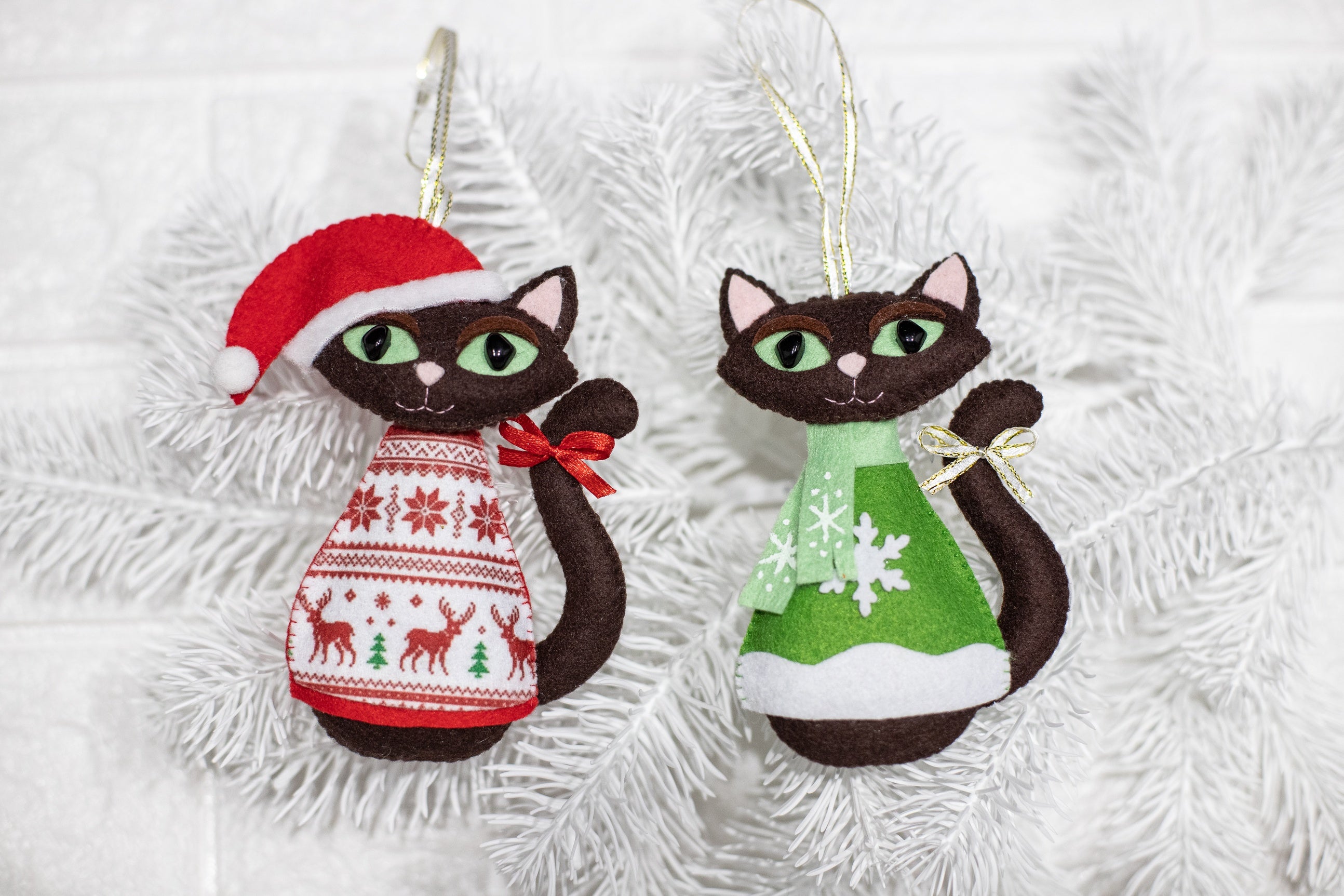 Brown felt cat Christmas ornament wearing a mint green scarf and green sweater with snowflake design, also brown cat wearing in red sweater and Santa hat. The cats has brown felt body,  black eyes, and a small gold or red bow on its tail. 