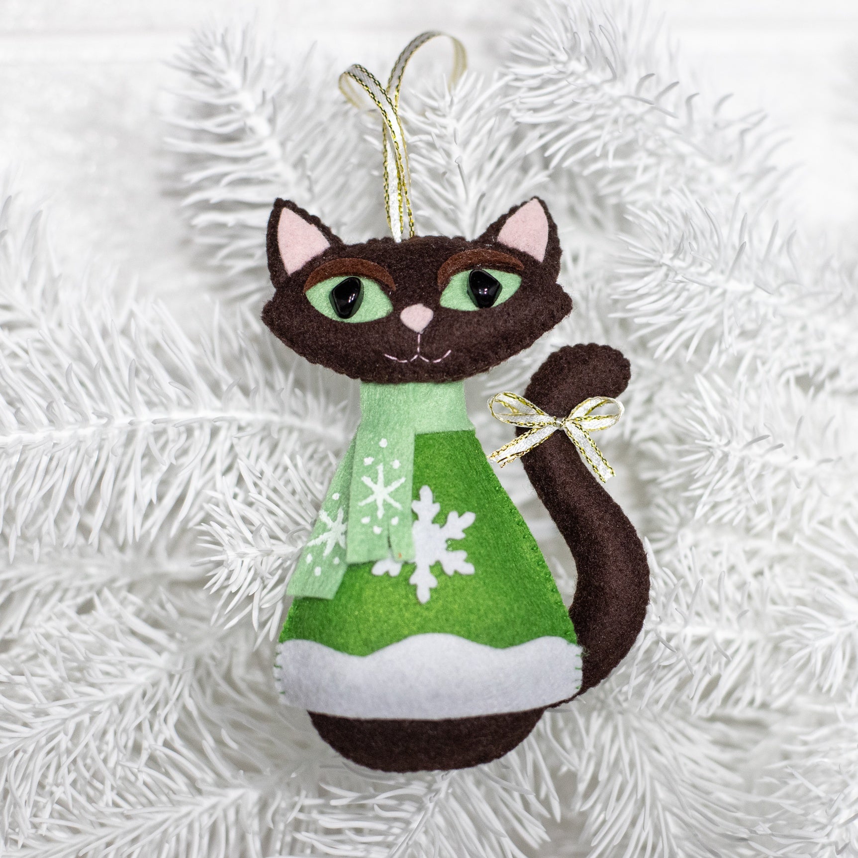 Brown felt cat Christmas ornament wearing a mint green scarf and green sweater with snowflake design. The cat has brown felt body,  black eyes, and a small gold bow on its tail. 