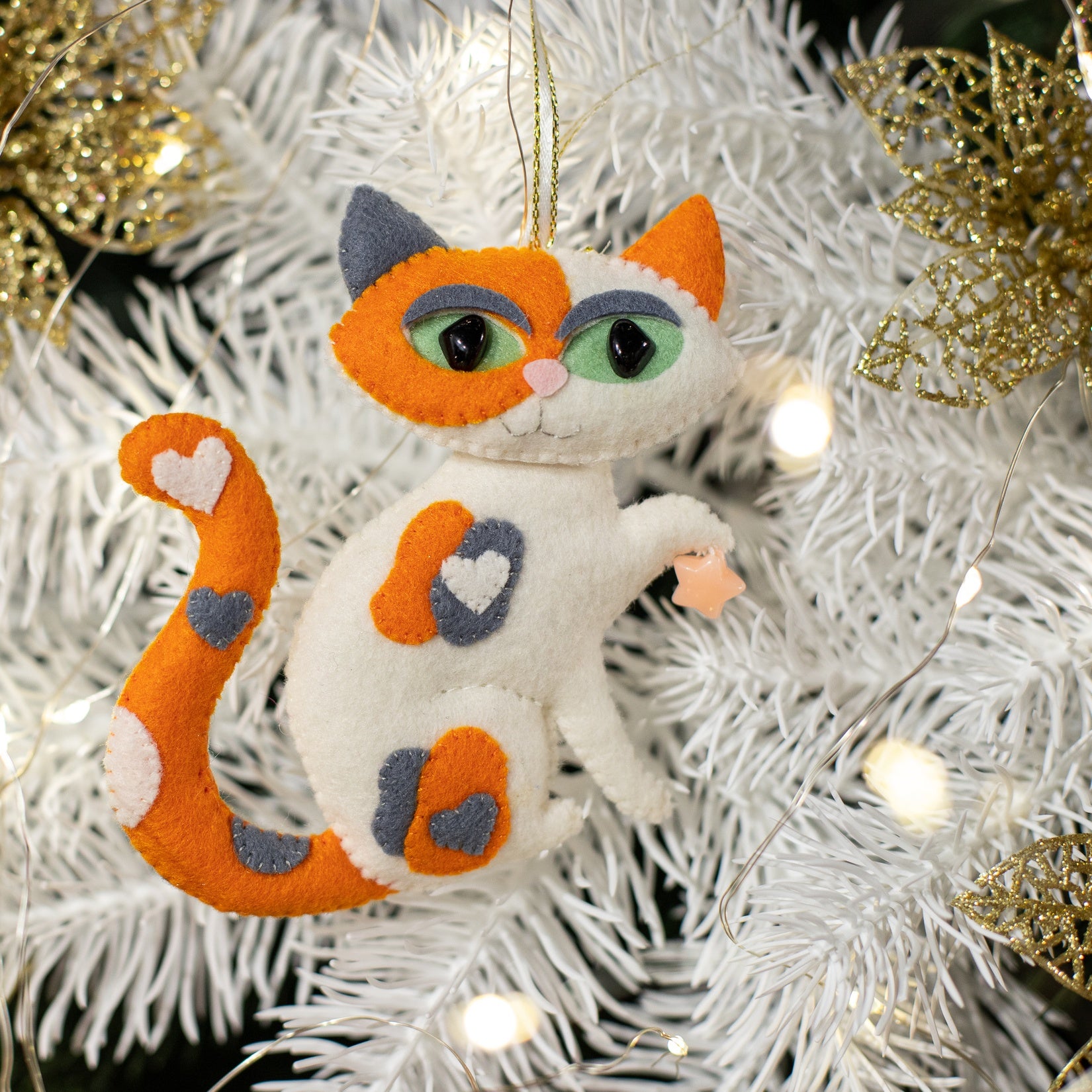 A white felt cat ornament with orange and grey patches, a small yellow bow, and a curled tail 