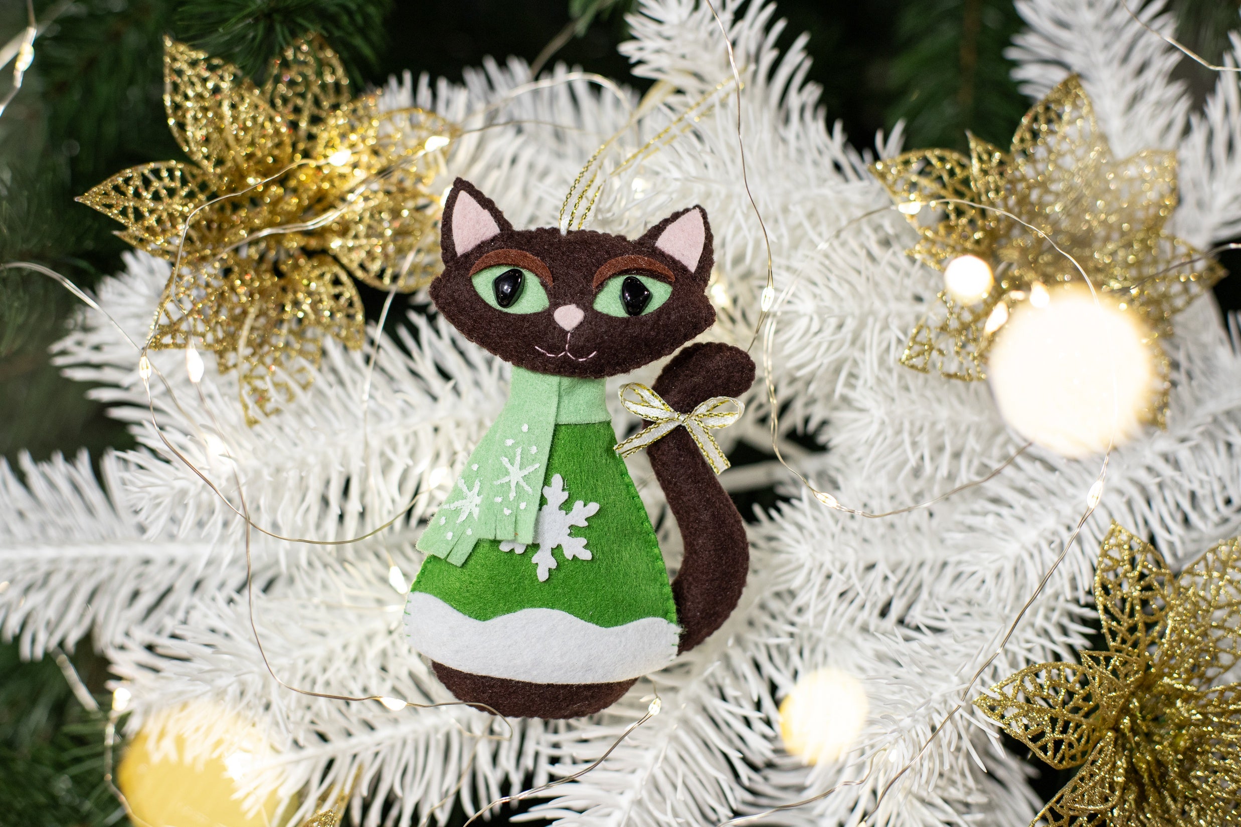 Brown felt cat Christmas ornament wearing a mint green scarf and green sweater with snowflake design. The cat has brown felt body,  black eyes, and a small gold bow on its tail. 