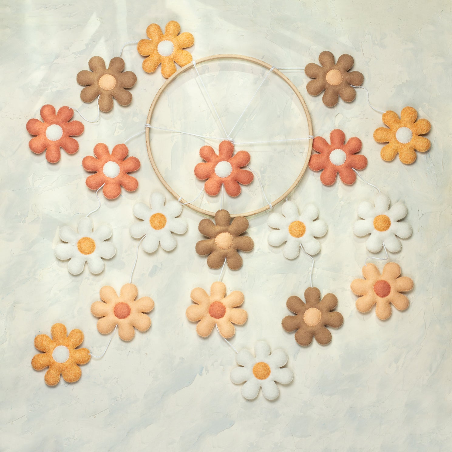 A handmade baby mobile hanging from a wooden arm, featuring several tiers of felt daisies in shades of terracotta, peach, tan, and white suspended from a light wooden hoop.