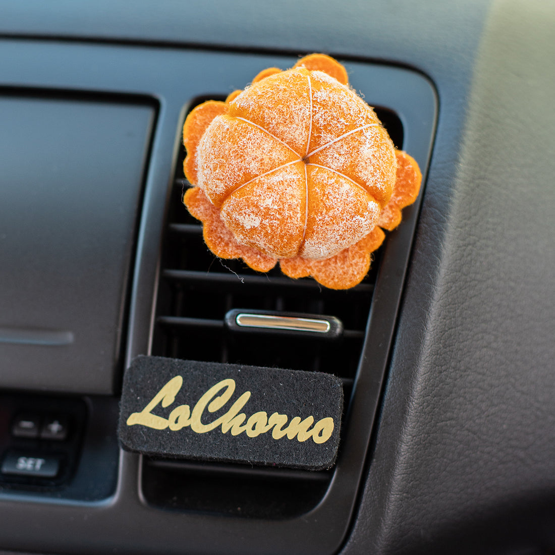 A handmade needle-felted mandarin fruit air freshener is clipped onto a black car air vent. Below the mandarin, a black felt tag with the word 'LoChorno' in gold script is clipped to the vent, customizable with any message or name.