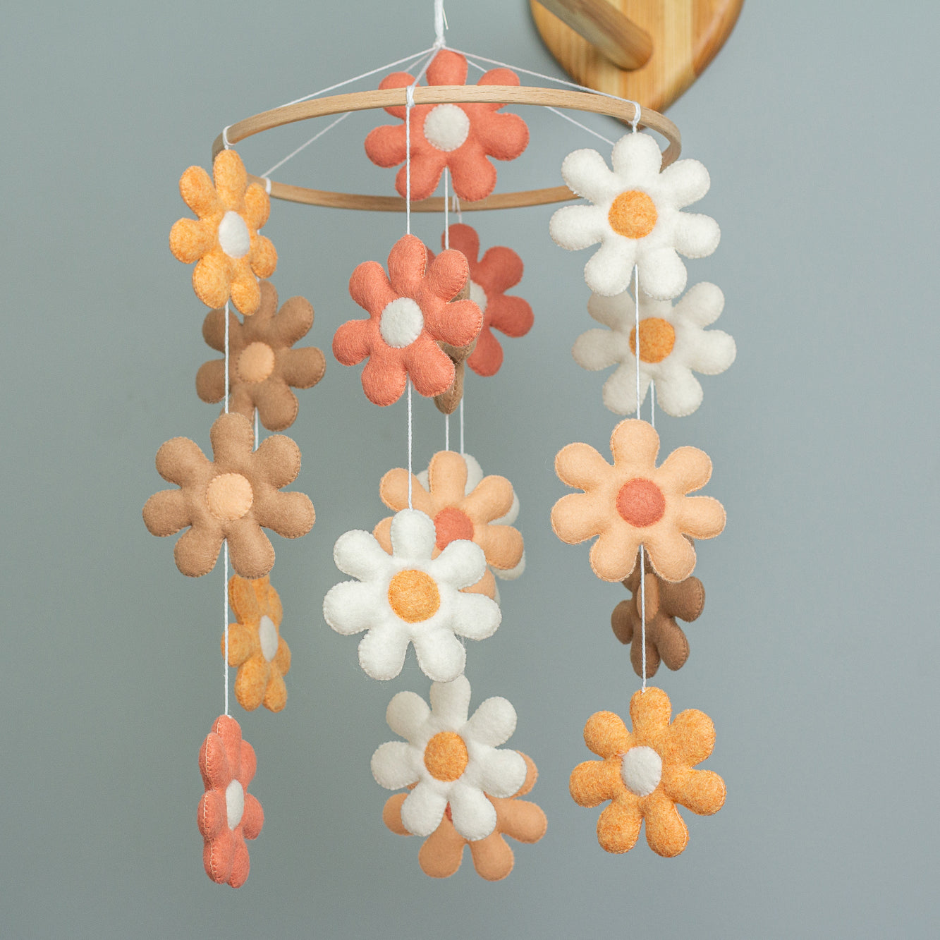A handmade baby mobile hanging from a wooden arm, featuring several tiers of felt daisies in shades of terracotta, peach, tan, and white suspended from a light wooden hoop.