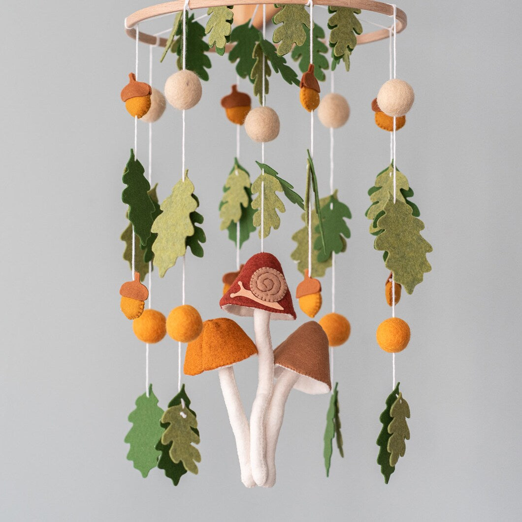 A handcrafted baby mobile featuring three large felt mushrooms in red, orange, and brown hanging in the center, with a small snail on one. Surrounding them are green oak leaves, orange acorns, and felt balls suspended from a wooden hoop.