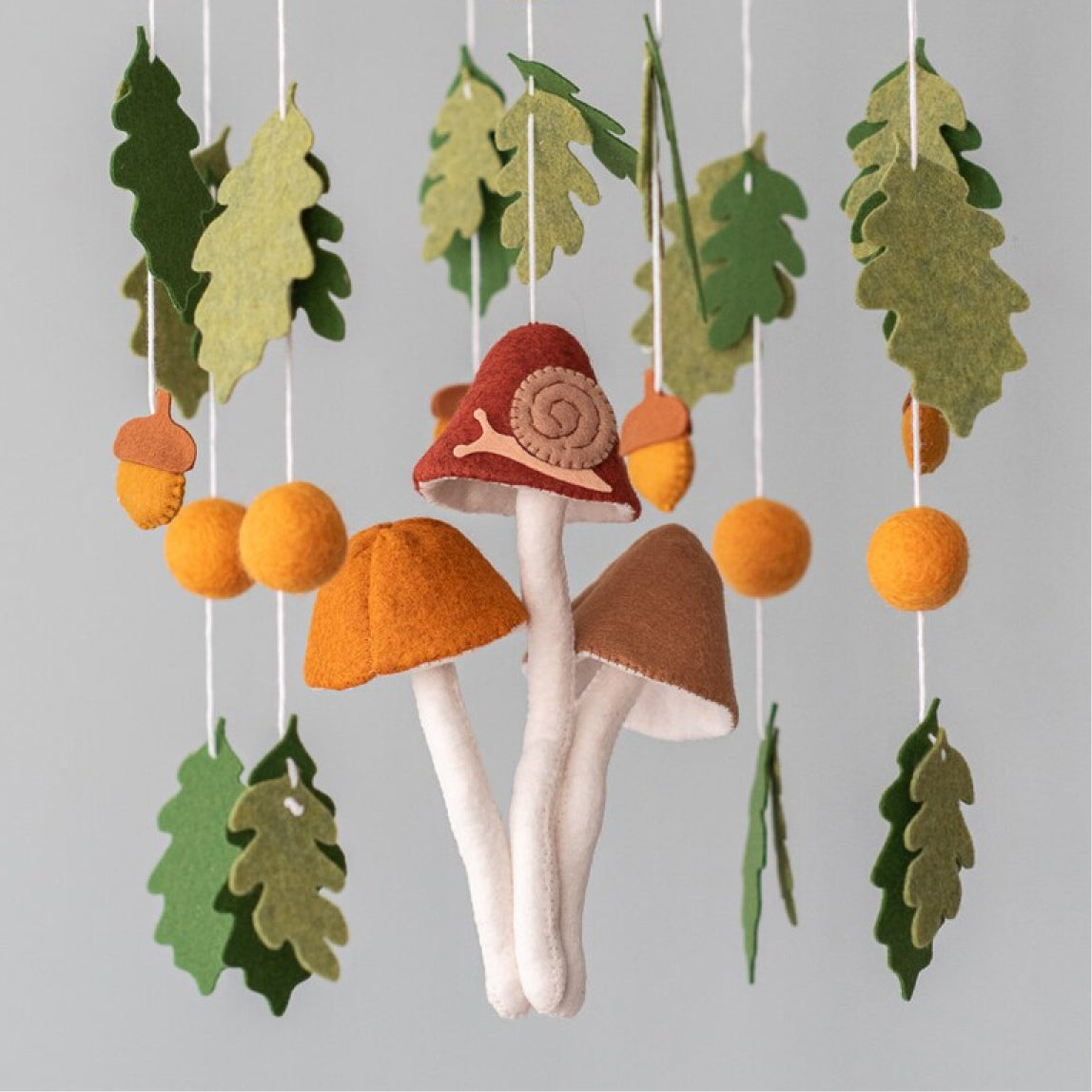 A handcrafted baby mobile featuring three large felt mushrooms in red, orange, and brown hanging in the center, with a small snail on one. Surrounding them are green oak leaves, orange acorns, and felt balls suspended from a wooden hoop.