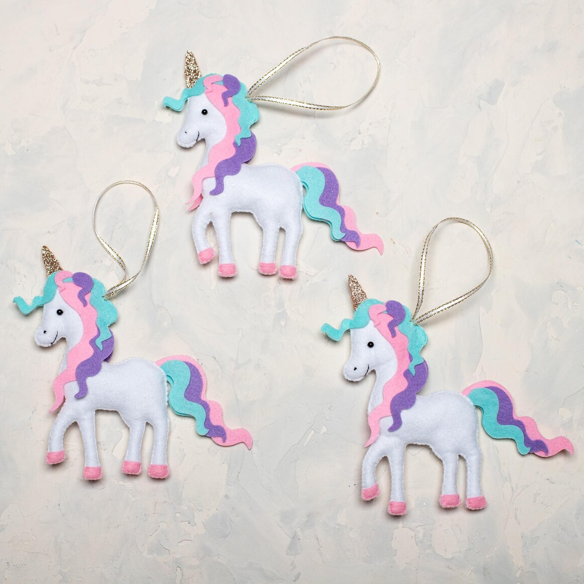 Three handmade white felt unicorn ornament with a wavy pastel mane and tail in pink, purple, and teal, a sparkly gold horn, and pink hooves, with a gold loop for hanging.